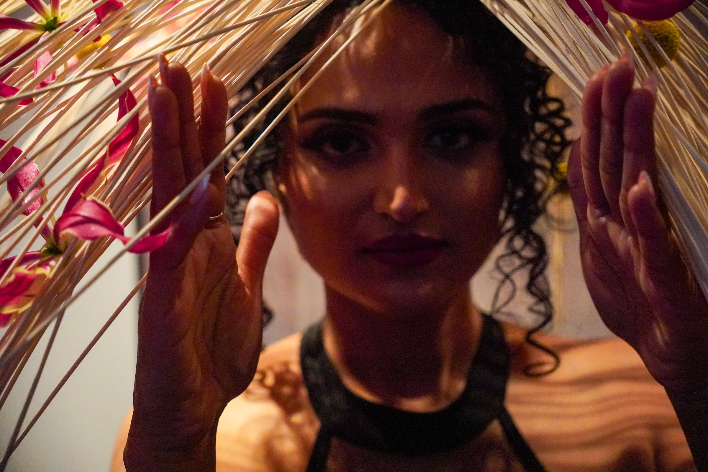 A face framed by radiating pale strands and pink flowers, with hands raised on either side.