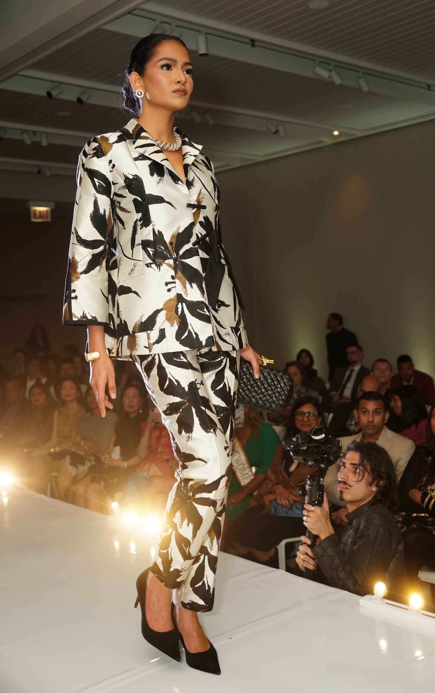 A model in a light jacket and trousers with dark leaf-like patterns walks past seated guests.