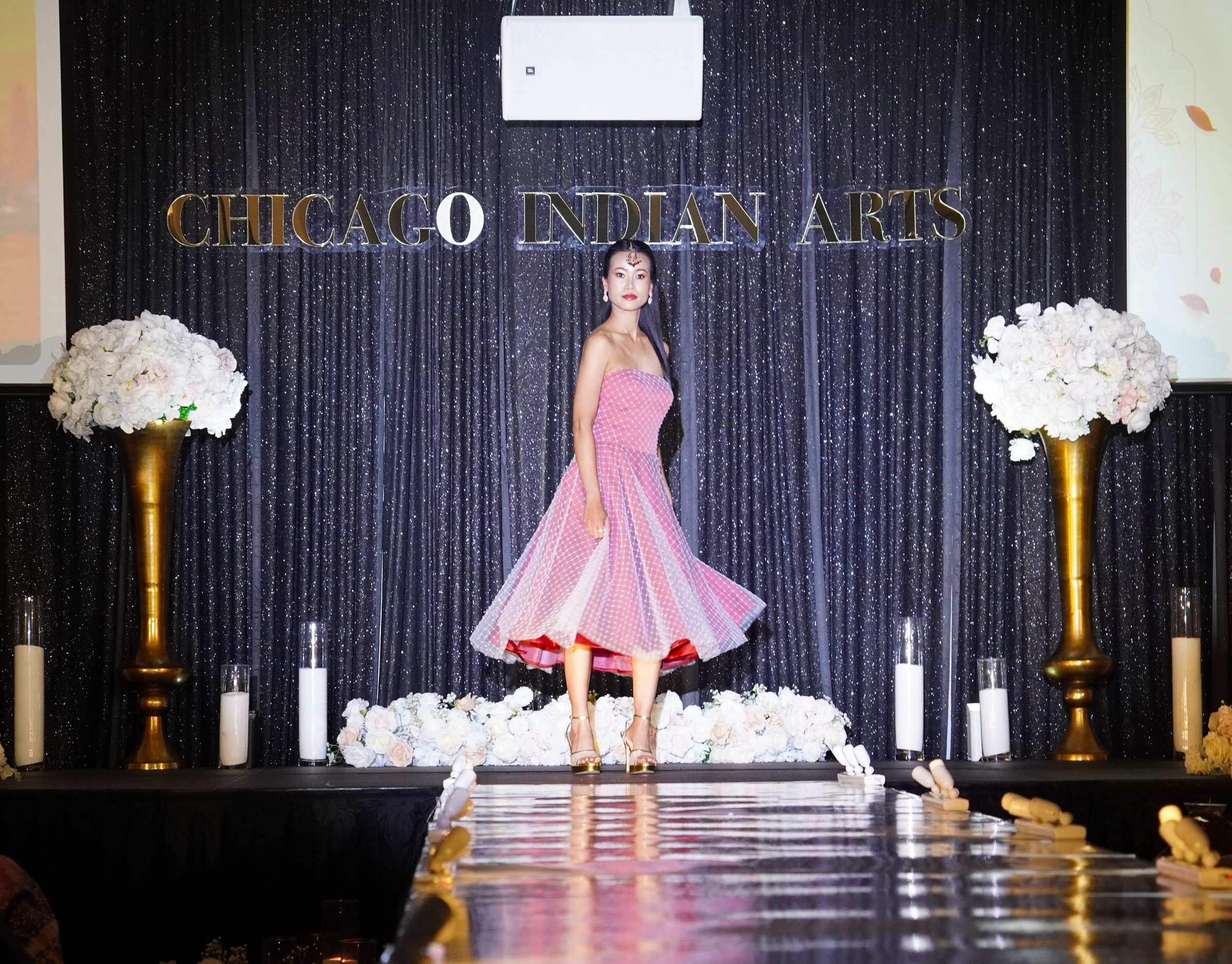 A model turns in a pink dress at the end of a reflective runway beneath Chicago Indian Arts lettering.